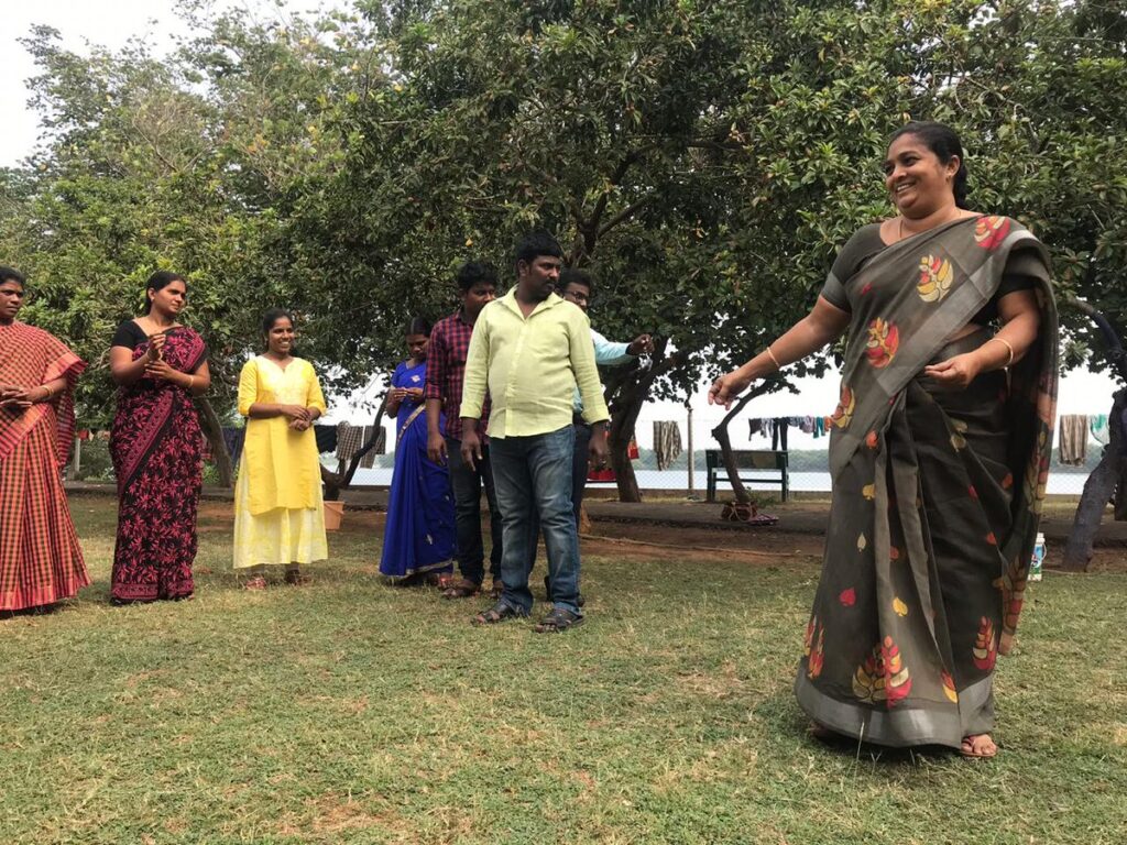 Smiling woman performing in a play. She is wearing a brown and orange patterned sari and she has long dark hair which is tied back. She has her arms outstretched almost as if she is dancing. Behind her a row of her colleagues look on. They are outside in a grassy area under some trees.