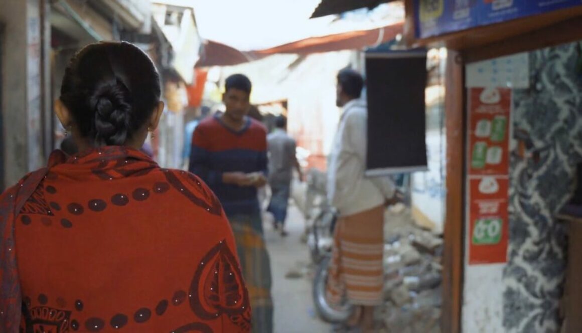 A woman in an orange top with dark, long hair which is tied back is seen from behind. She is in the foreground and in the background you can see she is walking down a narrow alley in an informal settlement. She is walking towards shops and three men in the distance who are walking around.