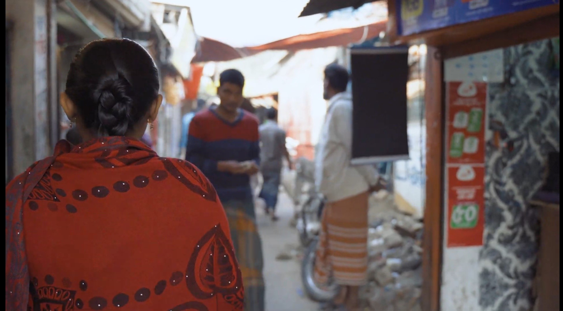 A woman in an orange top with dark, long hair which is tied back is seen from behind. She is in the foreground and in the background you can see she is walking down a narrow alley in an informal settlement. She is walking towards shops and three men in the distance who are walking around.
