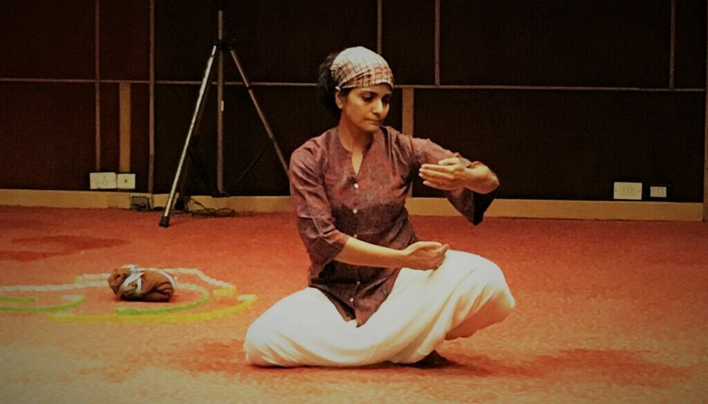 A woman sits cross legged on the floor. She is wearing white bottoms and an orange top. Her hair is tied back in a short scarf. She holds her arms out in front of her in a graceful dance move. She looks down to her fingertips. Her eyes are lowered. The carpet underneath her is an orange colour and behind her is dark wood paneling.