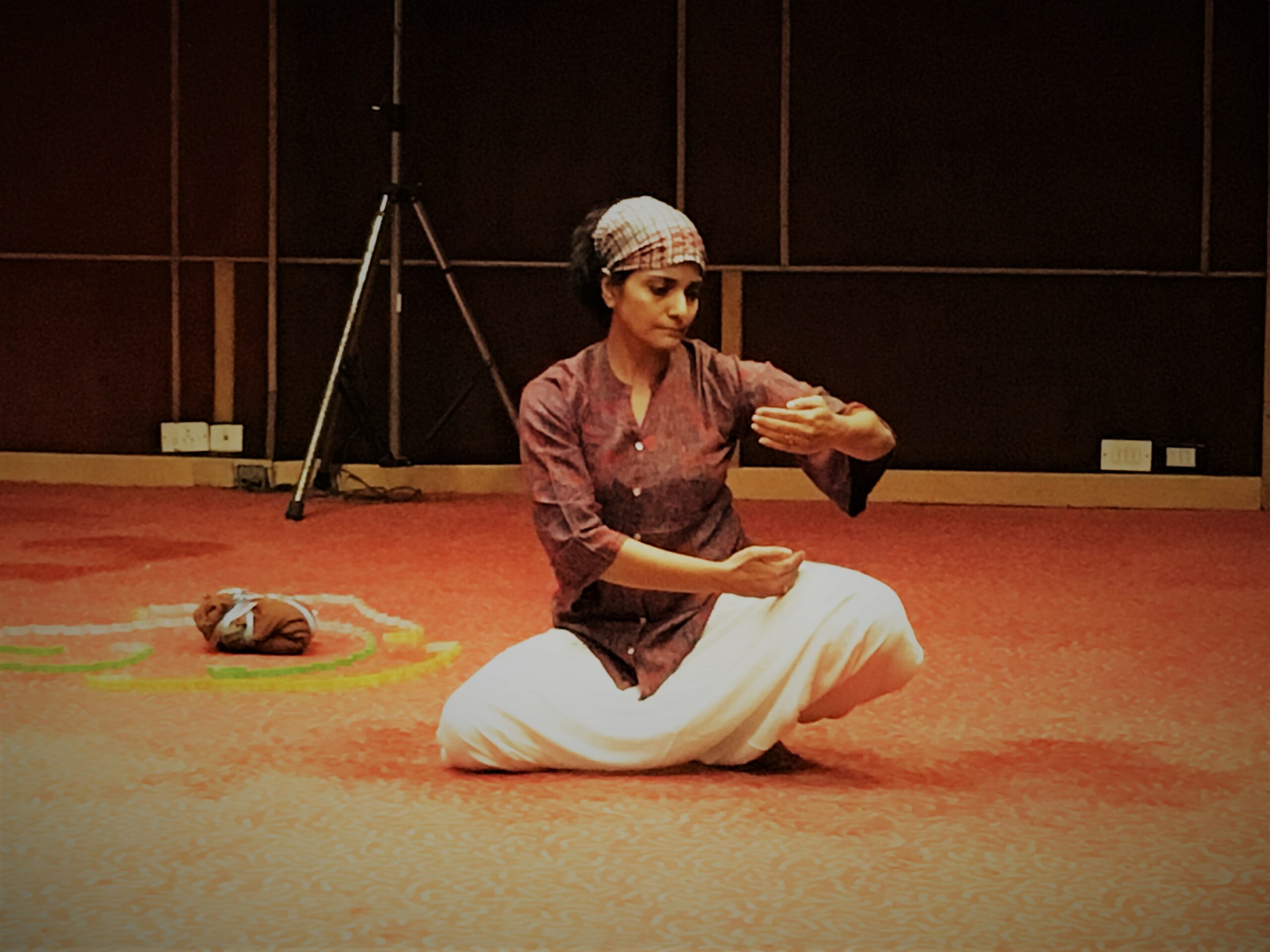 A woman sits cross legged on the floor. She is wearing white bottoms and an orange top. Her hair is tied back in a short scarf. She holds her arms out in front of her in a graceful dance move. She looks down to her fingertips. Her eyes are lowered. The carpet underneath her is an orange colour and behind her is dark wood paneling.
