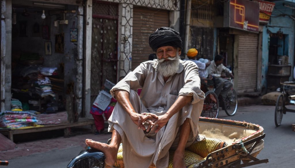 A rickshaw owner with an insecure livelihood sits cross legged facing the camera. He is older and he has a long white beard