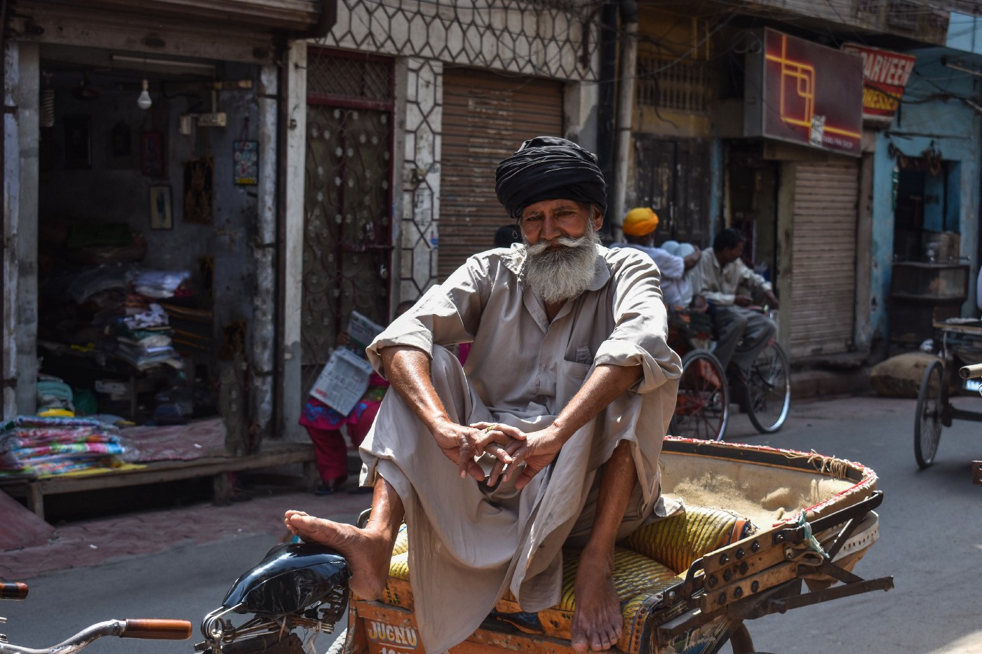 A rickshaw owner with an insecure livelihood sits cross legged facing the camera. He is older and he has a long white beard