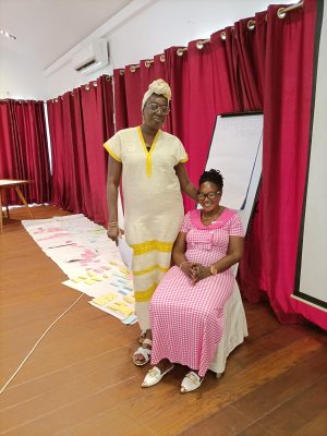 Colleagues and co researchers at the ARISE AGM, both women, one standing in a yellow dress and the other seated in a pink dress, inside the AGM venue